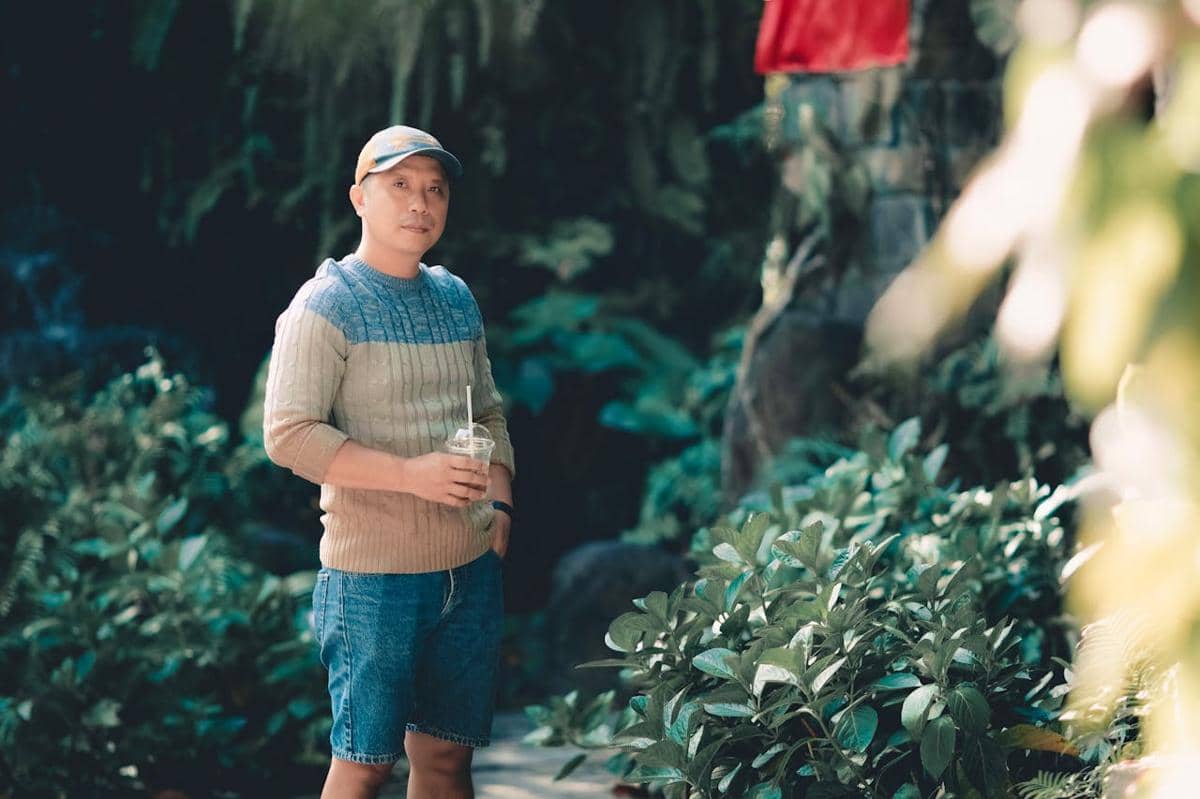 Pria mengenakan sweater crewneck dan celana pendek denim sambil memegang minuman di area taman dengan latar pepohonan hijau.