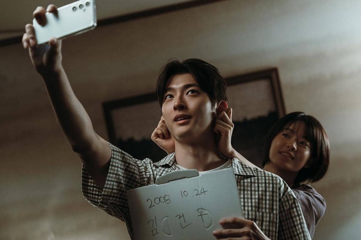 Dua orang muda berpose untuk swafoto, salah satunya memegang kertas bertuliskan tanggal dan nama dalam adegan serial If Wishes Could Kill.