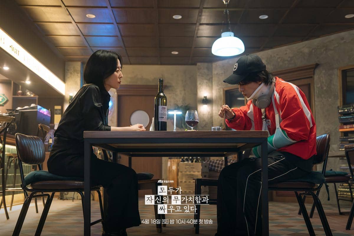 Dua orang duduk berhadapan di meja restoran dengan suasana hangat, satu mengenakan pakaian hitam dan lainnya jaket merah serta penyangga tangan.