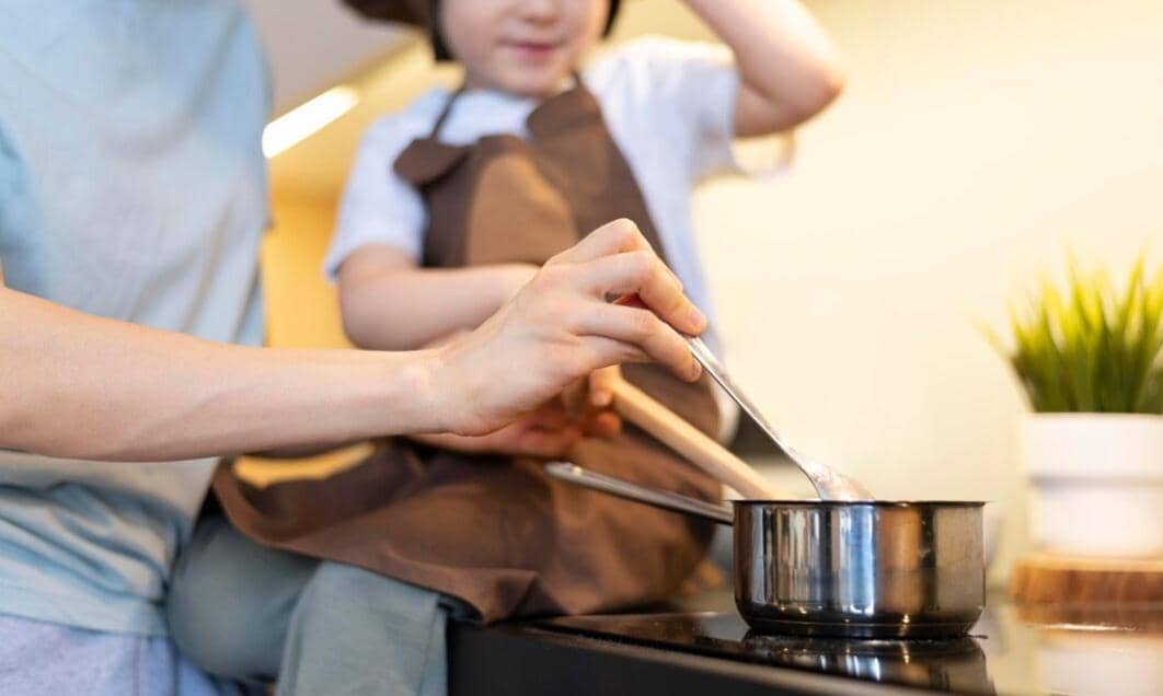 Seorang anak kecil mengenakan celemek cokelat belajar memasak bersama orang dewasa yang sedang mengaduk bahan di panci di dapur.