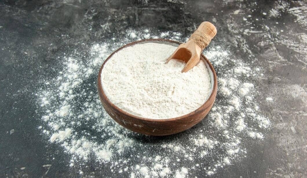 Mangkok berisi tepung maizena putih dengan sendok kayu kecil di atas meja gelap yang tertabur tepung di sekitarnya.
