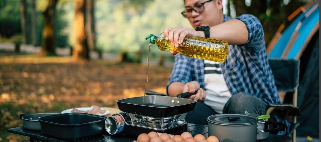 Seseorang menuang minyak dari botol plastik ke wajan di atas kompor portabel saat memasak di area luar ruangan dengan latar pepohonan.