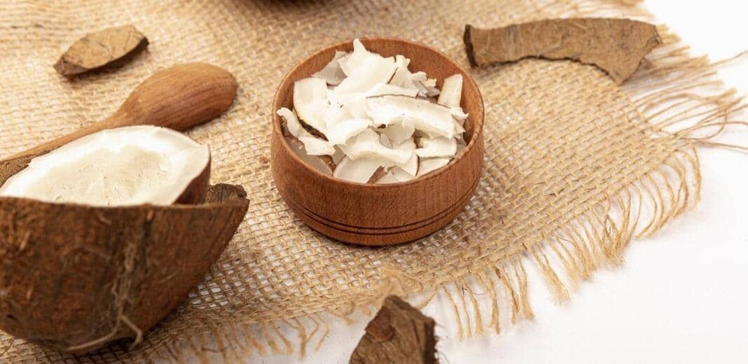 Potongan ampas kelapa putih dalam mangkuk kayu diletakkan di atas kain goni bersama tempurung kelapa dan sendok kayu.