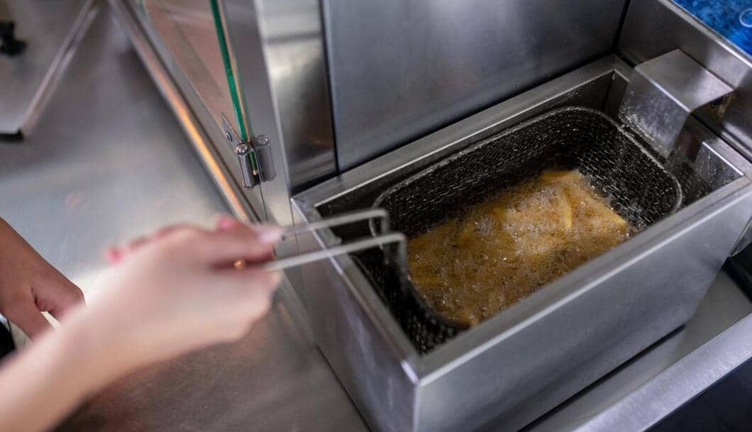 Seseorang menggoreng makanan menggunakan deep fryer stainless steel dengan minyak panas yang sedang mendidih di dapur komersial.
