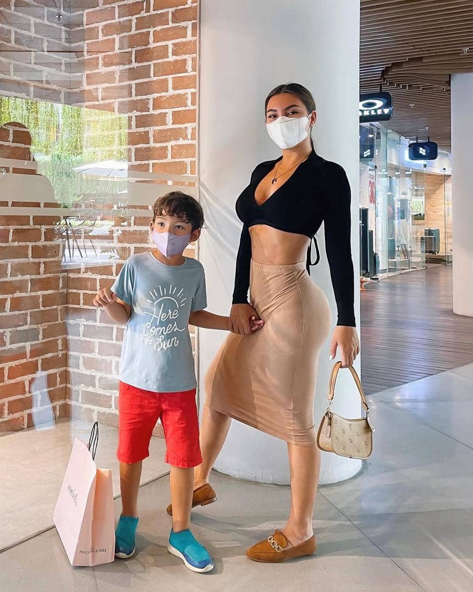 Seorang wanita mengenakan crop top hitam dan rok midi krem berdiri di mal sambil menggandeng anak laki-laki kecil yang memakai kaus biru dan celana merah.