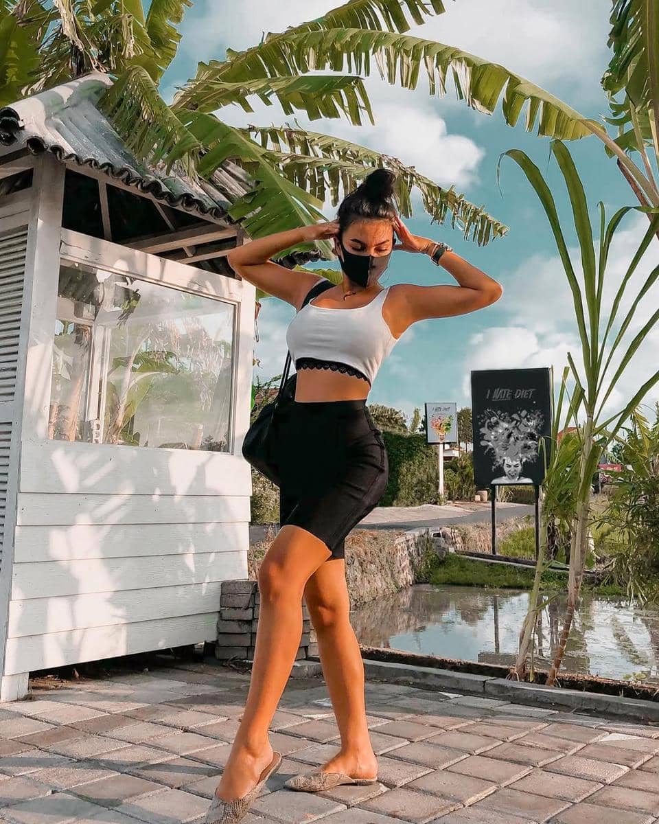 Seorang wanita mengenakan crop top putih dan rok hitam berpose di luar ruangan dengan latar pepohonan pisang dan langit cerah.