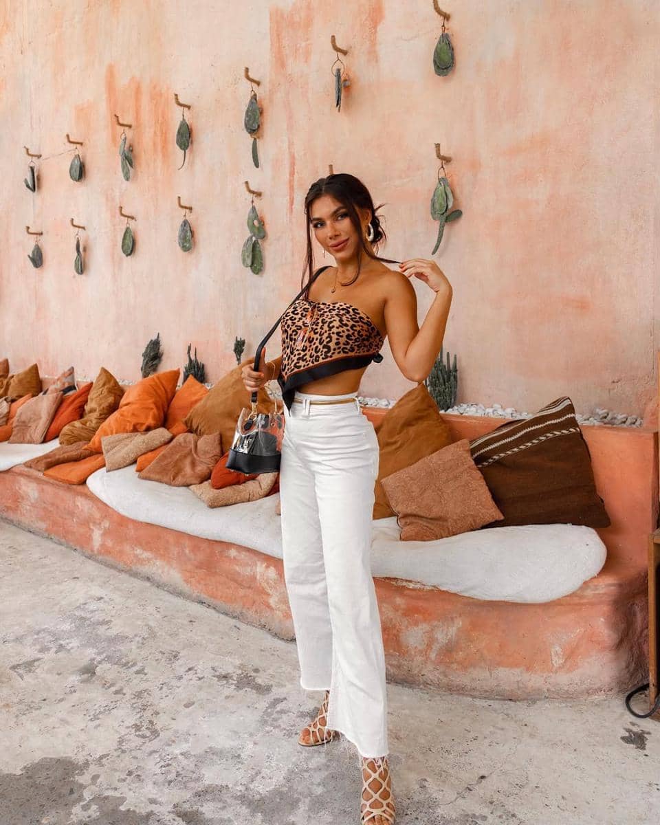 Seorang wanita berpose dengan crop top motif leopard dan celana panjang putih di depan dinding berwarna peach dengan dekorasi tanaman gantung.