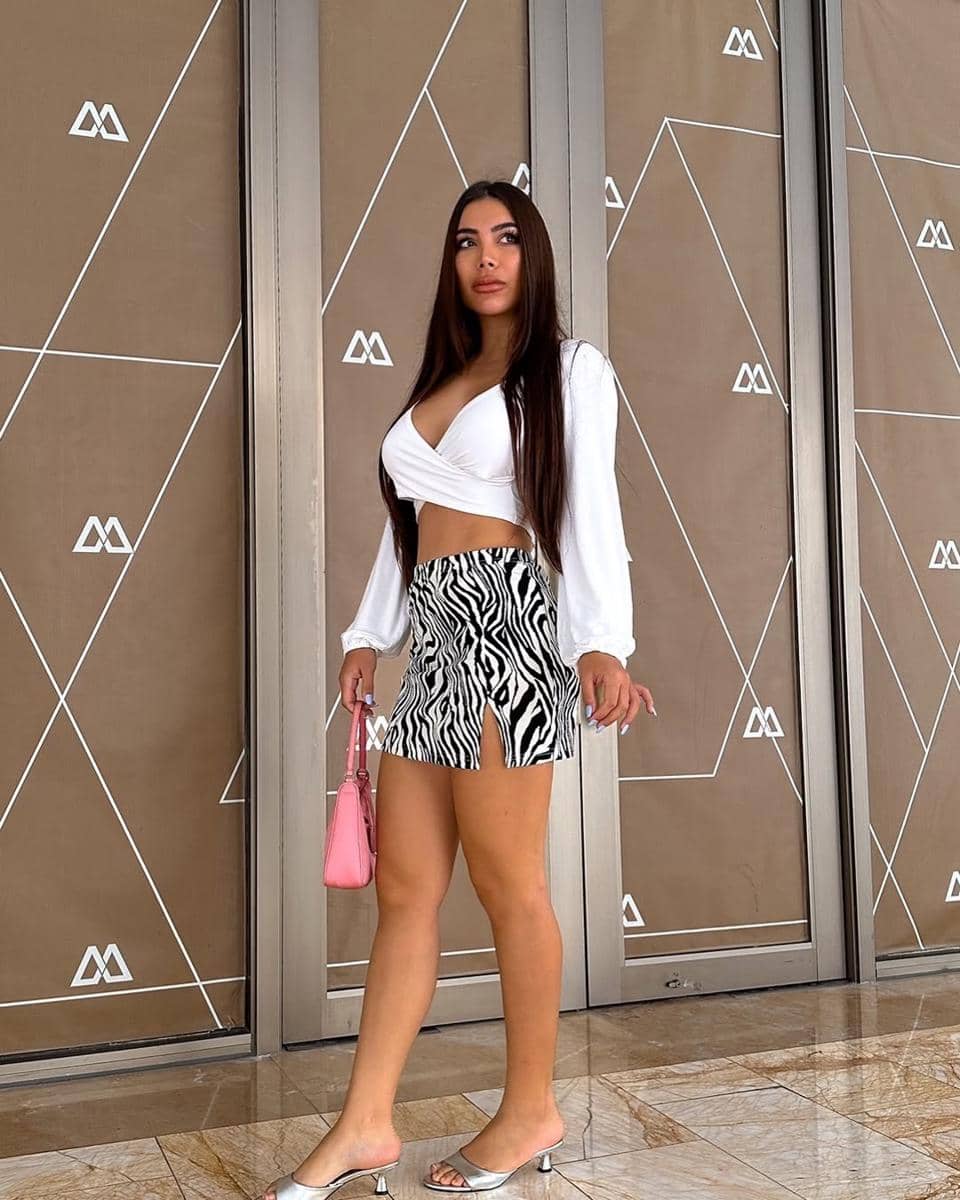 Seorang wanita mengenakan crop top putih dengan rok mini motif zebra, membawa tas kecil berwarna pink dan sandal hak rendah.