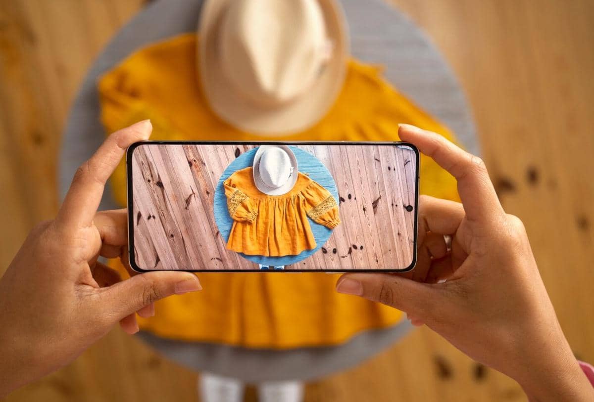 Seseorang memotret baju kuning dan topi putih di atas meja kayu menggunakan ponsel pintar dengan tampilan hasil foto di layar.