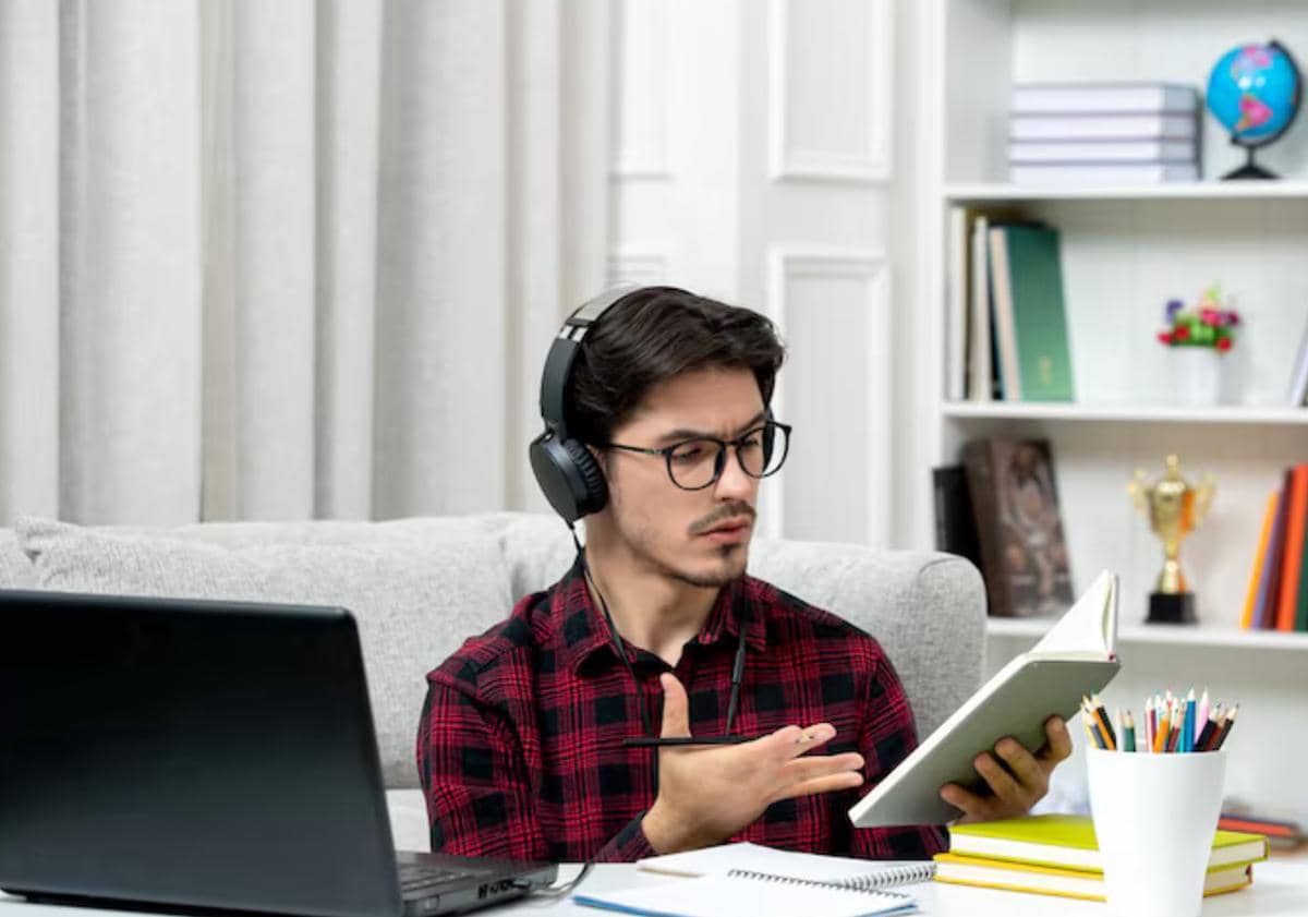 Seorang pria mengenakan headphone dan kemeja kotak-kotak merah sedang membaca buku di depan laptop di ruang belajar rumah.