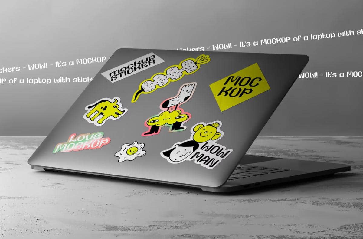 Laptop abu-abu dengan berbagai stiker ilustrasi berwarna cerah seperti karakter lucu, tulisan, dan bentuk unik di permukaannya.