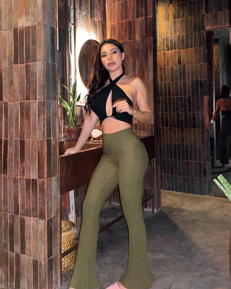 Seorang wanita mengenakan crop top hitam dan celana panjang hijau berdiri di ruangan berdinding bata dengan pencahayaan hangat.