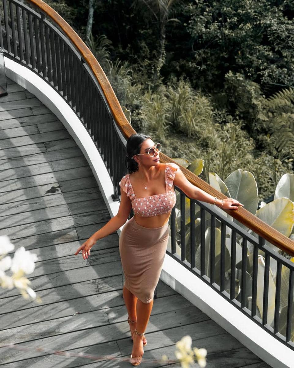 Seorang wanita mengenakan crop top bermotif dan rok midi berwarna netral berjalan di jembatan kayu dengan latar pepohonan hijau.