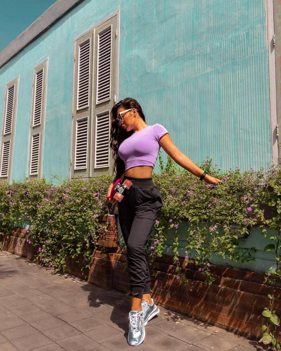 Seorang wanita mengenakan crop top ungu dan celana jogger hitam berpose di trotoar dengan latar dinding hijau dan tanaman berbunga ungu.