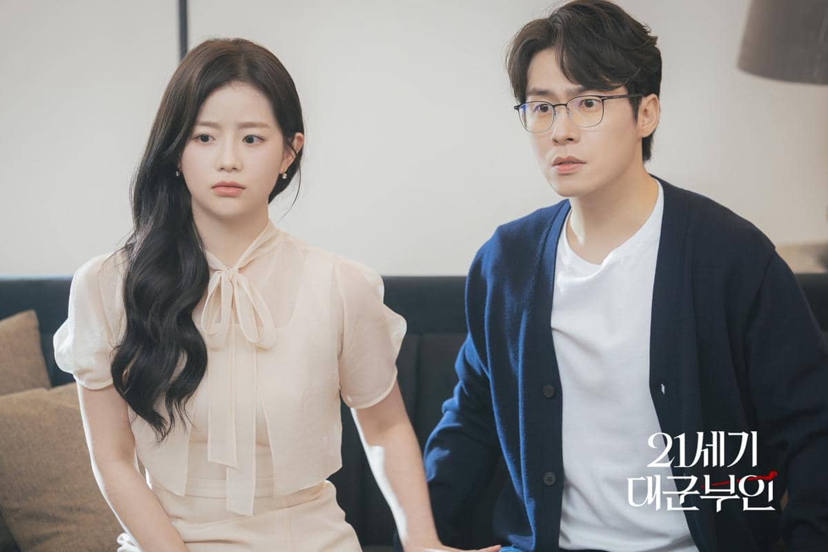 Dua pemeran drama Perfect Crown duduk berdampingan di sofa, menampilkan ekspresi serius dalam suasana ruangan yang tenang.