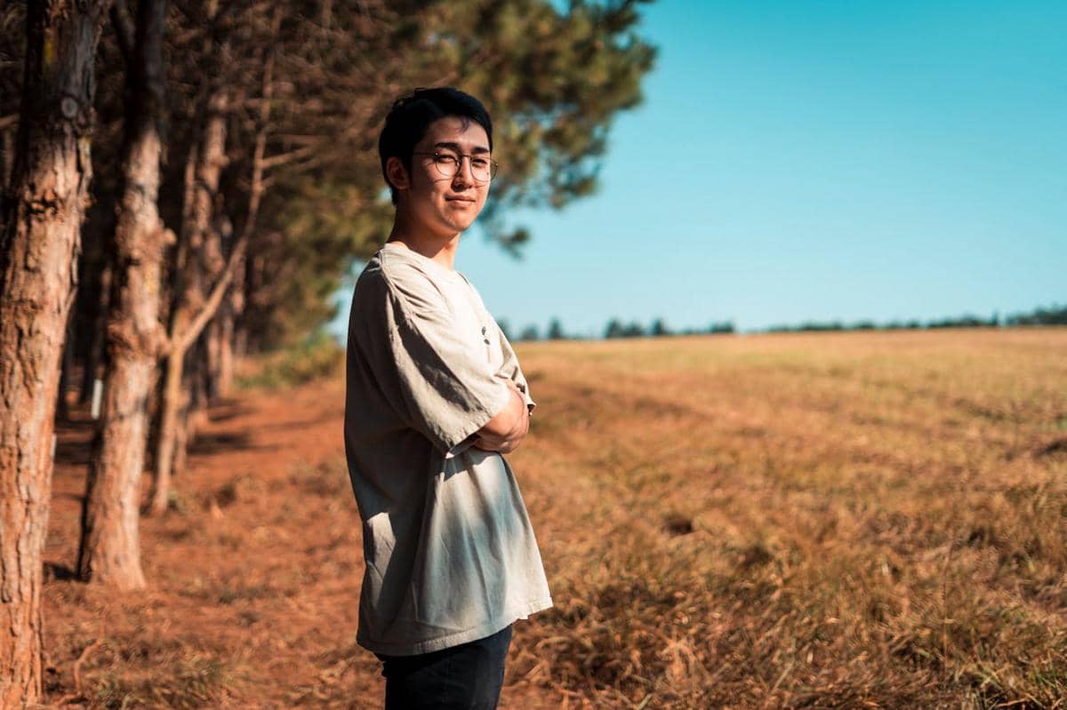 Pria berdiri dengan tenang di tepi hutan pinus, mengenakan kaus abu-abu dan kacamata di bawah langit cerah biru.