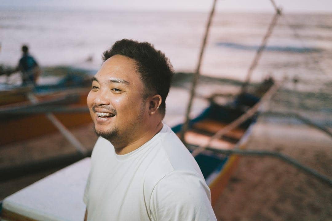 Pria tersenyum mengenakan kaus putih di tepi pantai dengan latar perahu tradisional dan laut saat senja.