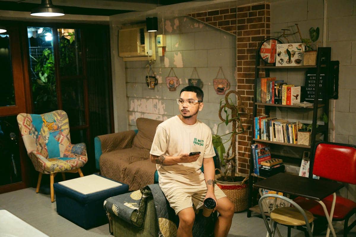 Seorang pria duduk di ruang tamu bergaya retro dengan rak buku, kursi warna-warni, dan pencahayaan hangat sambil memegang ponsel.