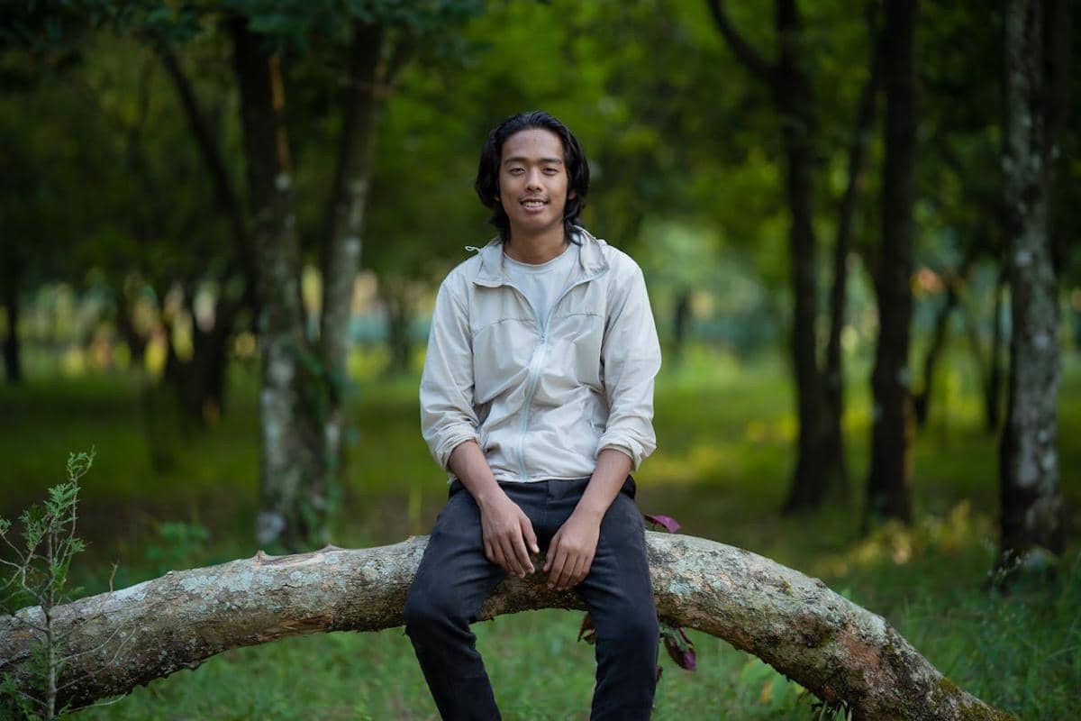 Seorang pria berjaket krem duduk santai di batang pohon tumbang di tengah hutan hijau dengan latar pepohonan rindang.