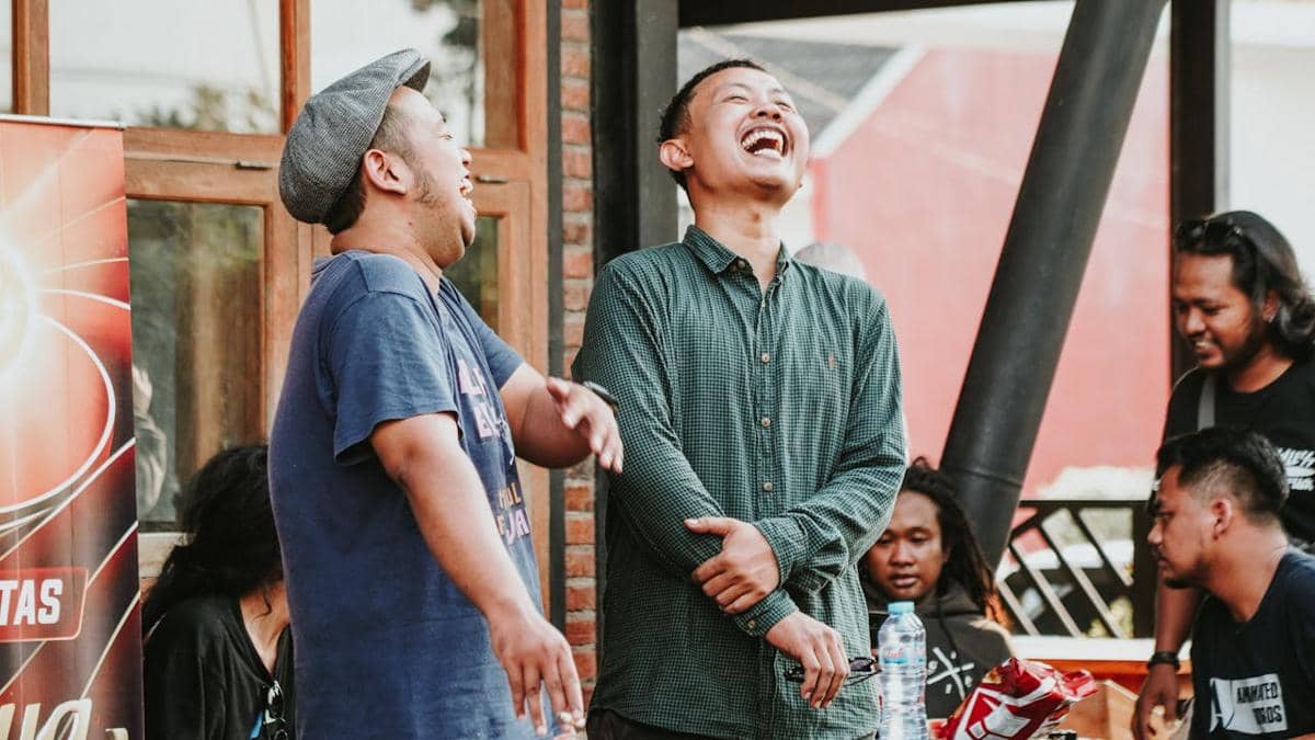 Sekelompok teman tertawa bersama di luar ruangan, dua pria tampak berbagi momen lucu sementara lainnya duduk di sekitar meja.