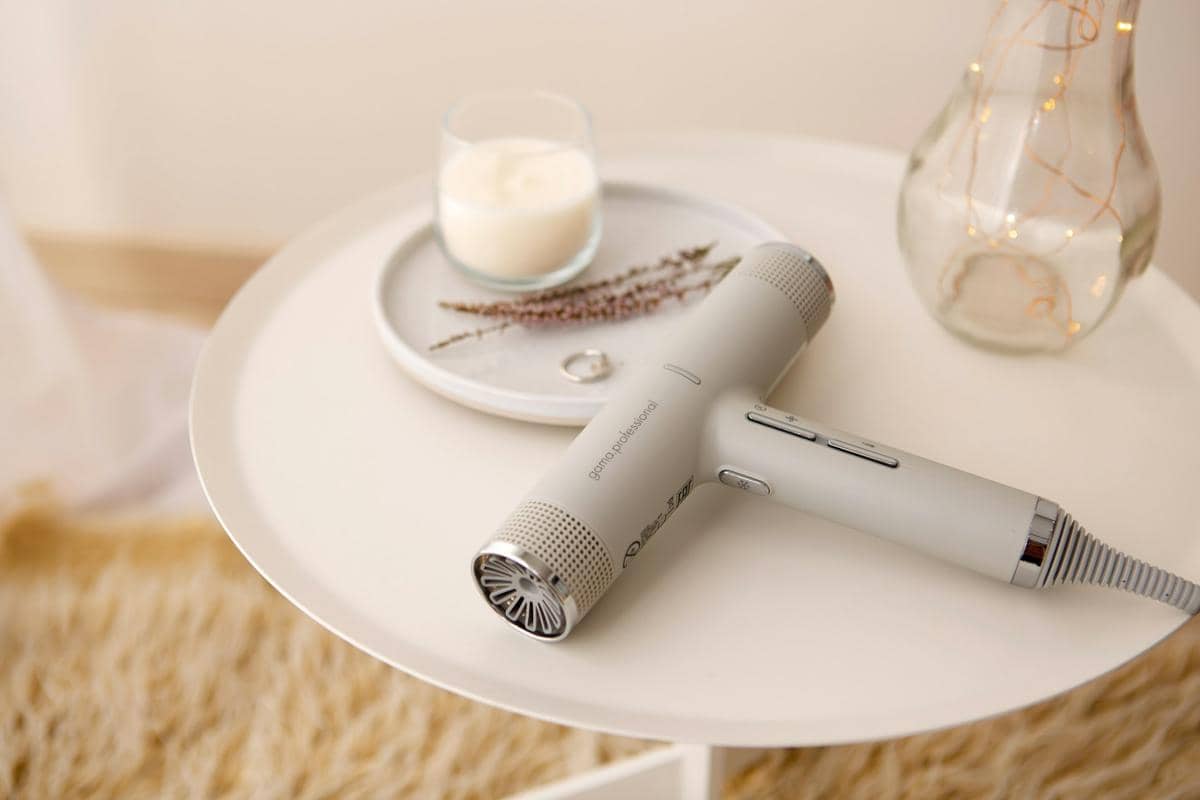 Hair dryer berwarna abu-abu muda terletak di atas meja bundar putih dengan lilin dan hiasan kering di sampingnya.