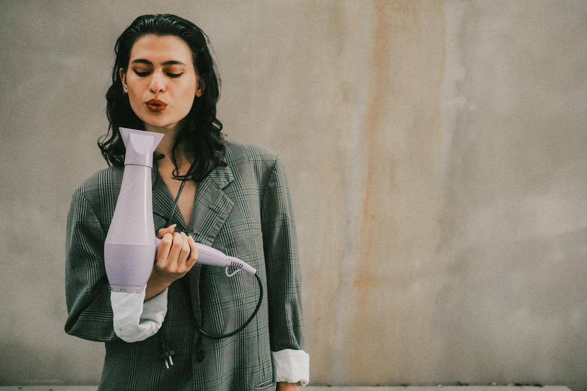 Seorang perempuan berambut hitam mengenakan jas kotak-kotak memegang hair dryer ungu muda di depan dinding berwarna netral.