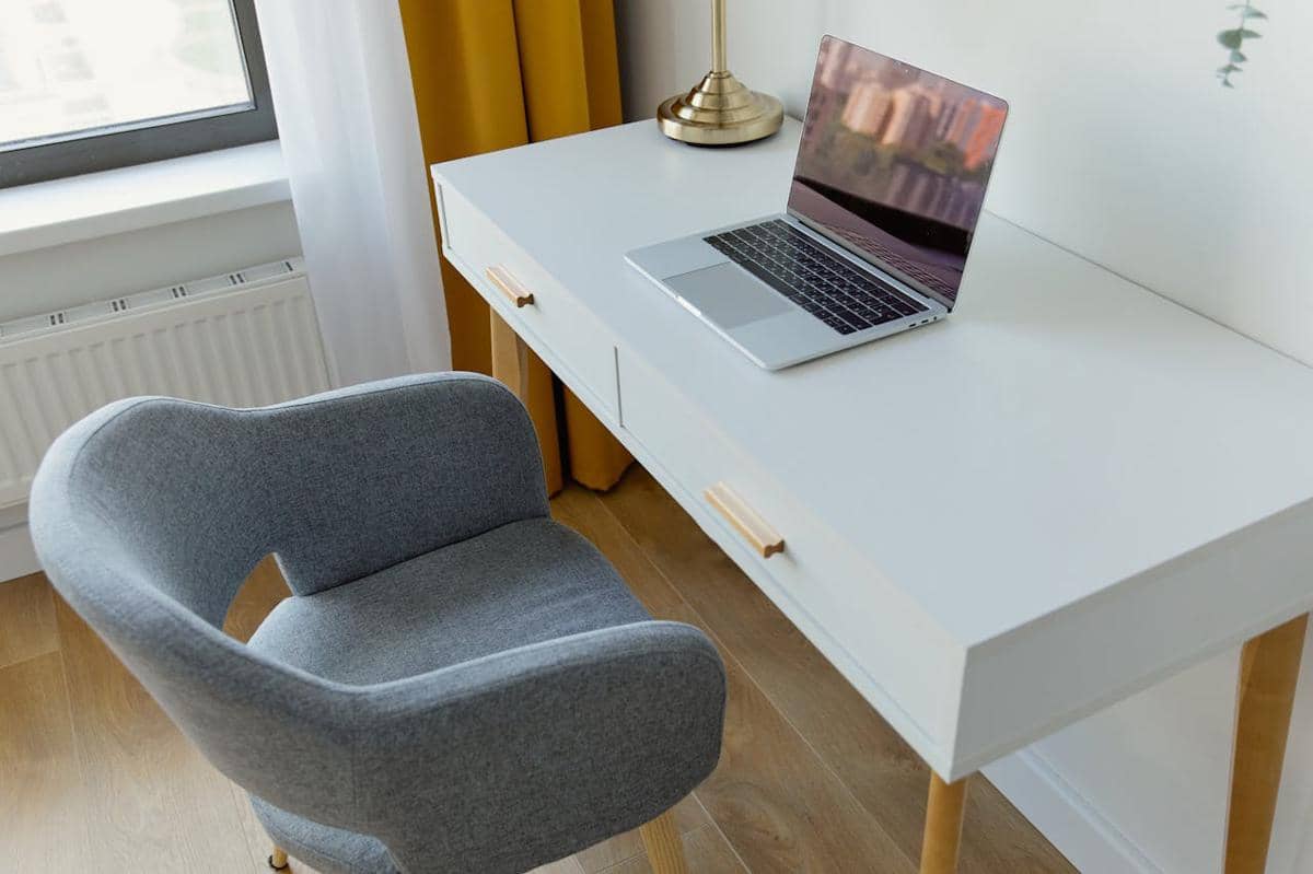 Meja belajar minimalis berwarna putih dengan laptop terbuka di atasnya, kursi abu-abu, dan tirai kuning di dekat jendela.