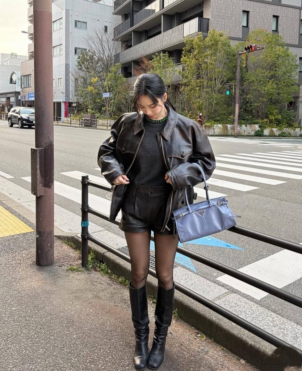 Seorang wanita berdiri di trotoar kota mengenakan jaket kulit hitam, celana pendek, dan sepatu bot tinggi sambil membawa tas biru.