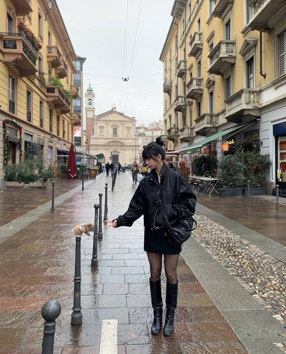 Seorang wanita berjaket hitam berdiri di jalan kota Eropa yang basah setelah hujan, menyentuh sarung tangan di tiang besi.