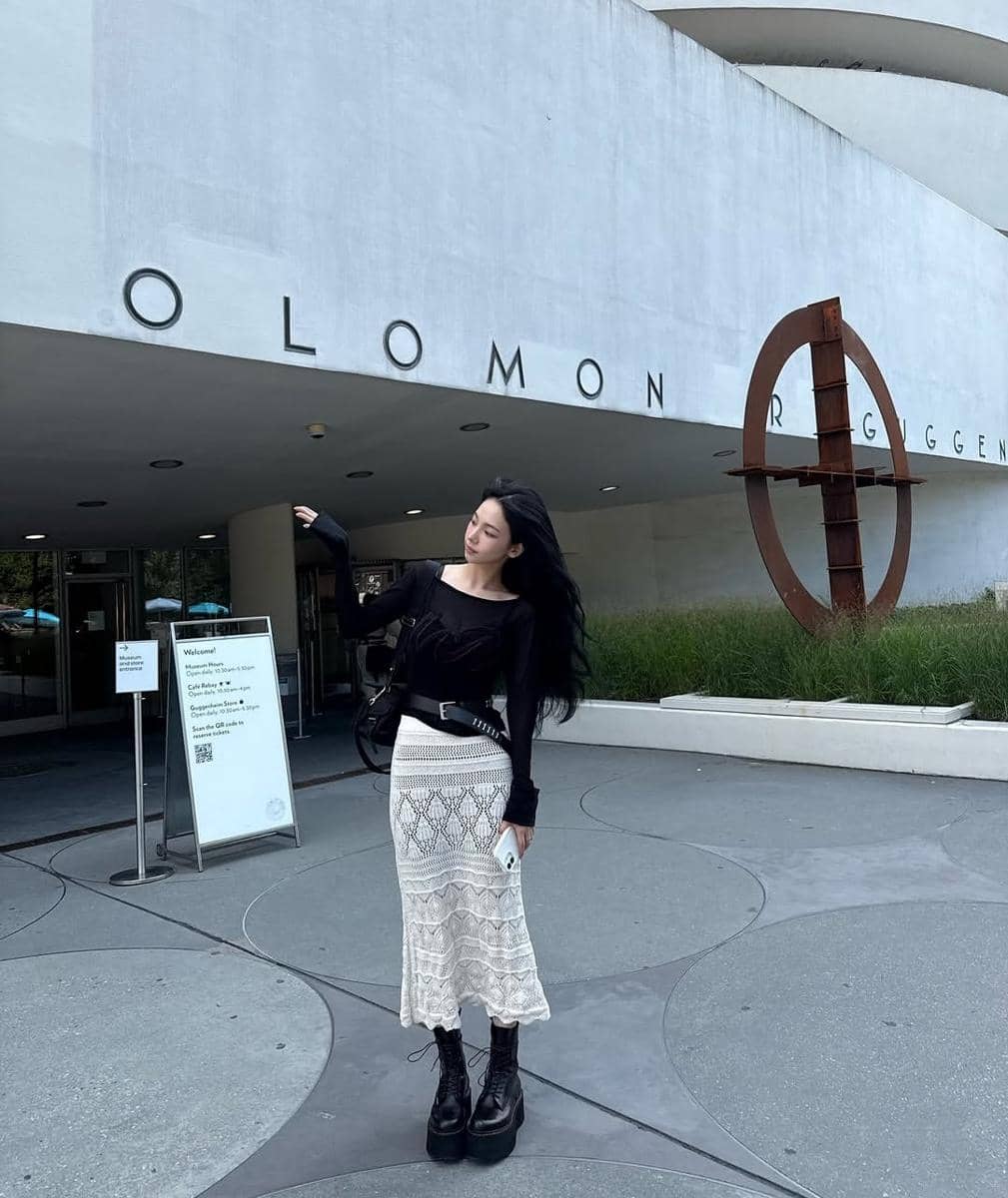 Seorang wanita berpose di depan bangunan museum dengan dinding putih melengkung dan tulisan besar di fasadnya.