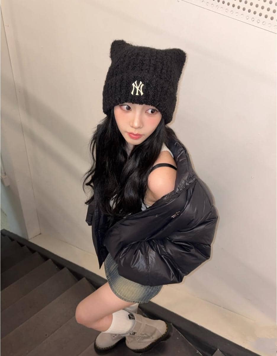 Seorang perempuan berpose di tangga mengenakan jaket hitam tebal, rok pendek, topi rajut hitam dengan logo NY, dan sepatu boots krem.