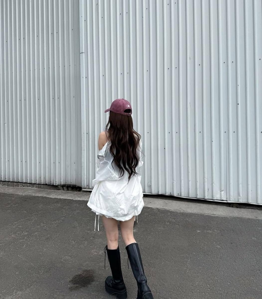 Seorang perempuan berambut panjang mengenakan jaket putih, topi merah marun, dan sepatu bot hitam berjalan di dekat dinding logam abu-abu.