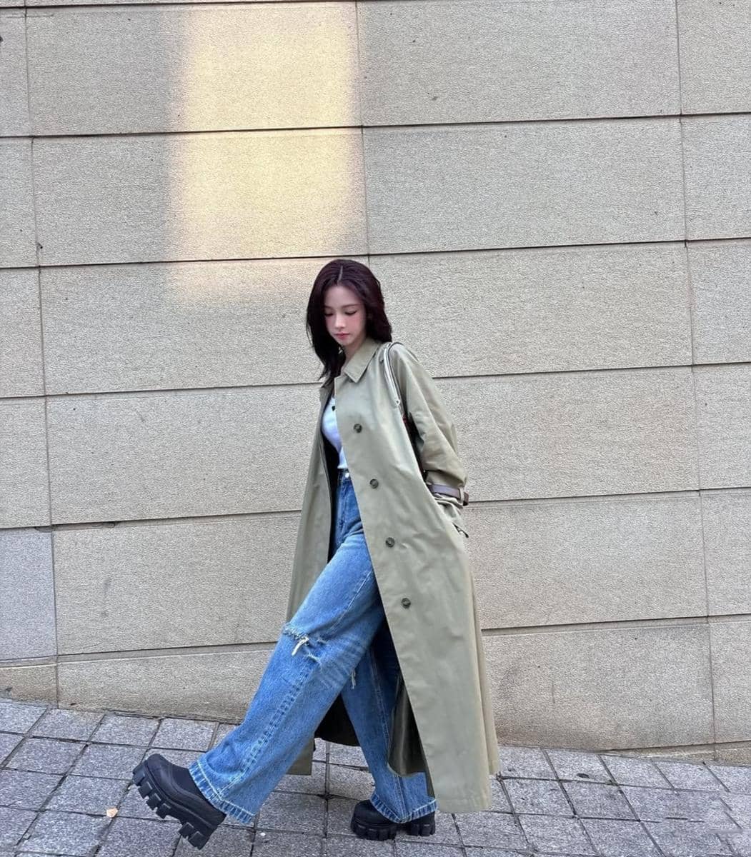 Seorang wanita mengenakan trench coat hijau zaitun panjang, celana jeans longgar, dan sepatu platform hitam berjalan di trotoar.