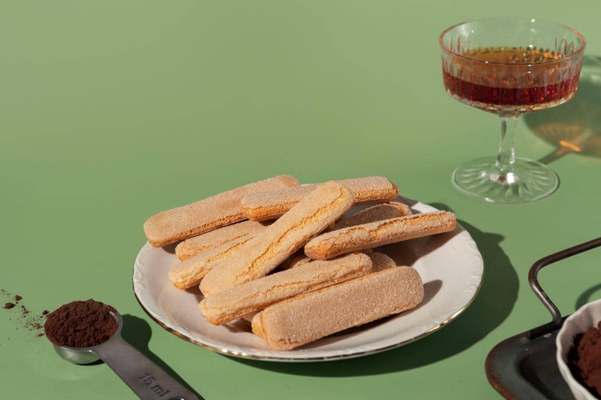 Beberapa biskuit ladyfinger berwarna keemasan tersusun di atas piring putih dengan latar meja hijau dan segelas minuman di sampingnya.