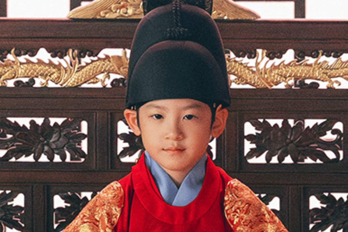 Anak laki-laki mengenakan hanbok kerajaan berwarna merah dan topi tradisional hitam duduk di depan latar ornamen kayu berukir emas.
