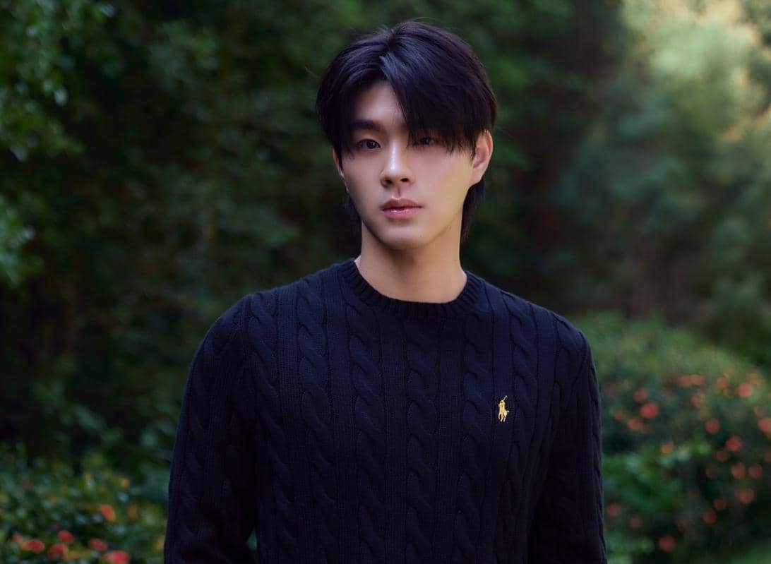 Seorang pria berambut hitam mengenakan sweter rajut hitam berdiri di luar ruangan dengan latar pepohonan hijau.