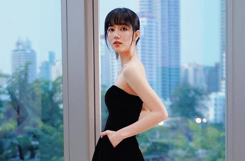 Seorang wanita berambut hitam mengenakan gaun hitam tanpa tali berdiri di depan jendela besar dengan latar gedung pencakar langit.