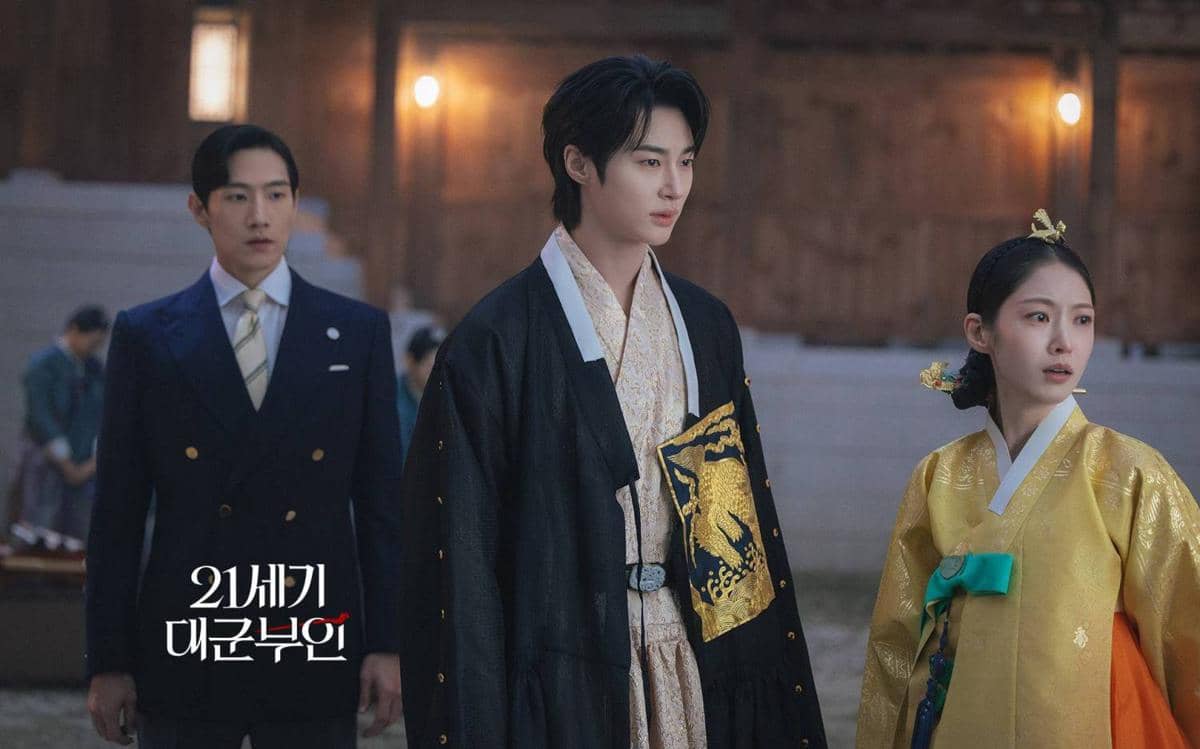 Tiga pemeran drama Perfect Crown mengenakan busana tradisional dan modern Korea berdiri di halaman istana pada malam hari.