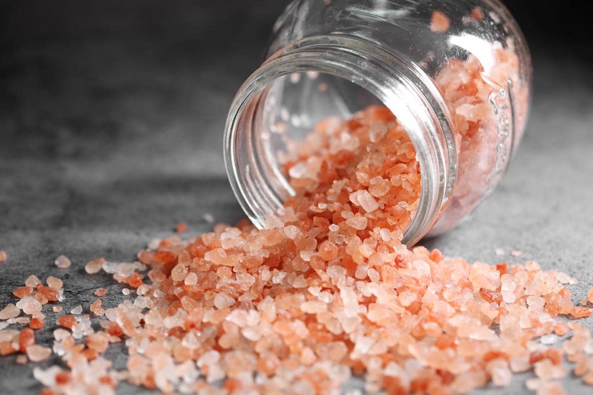 Butiran garam Himalaya berwarna merah muda keluar dari toples kaca di atas permukaan abu-abu dengan pencahayaan lembut.