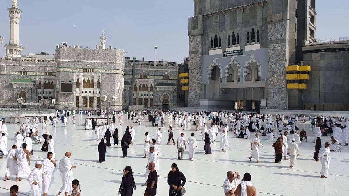 Sejumlah jemaah mengenakan pakaian ihram berjalan kaki di area Masjidil Haram dengan latar bangunan megah dan menara tinggi.