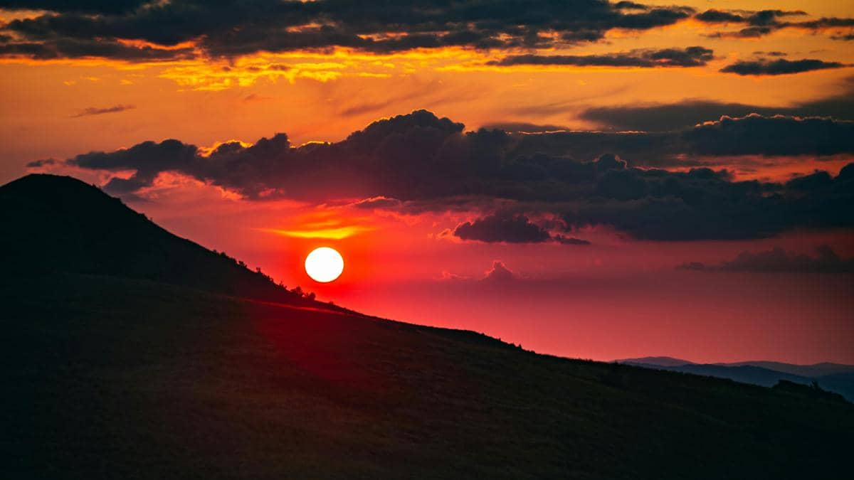 Pemandangan matahari terbenam di balik perbukitan dengan langit berwarna jingga, merah, dan ungu yang dipenuhi awan gelap.