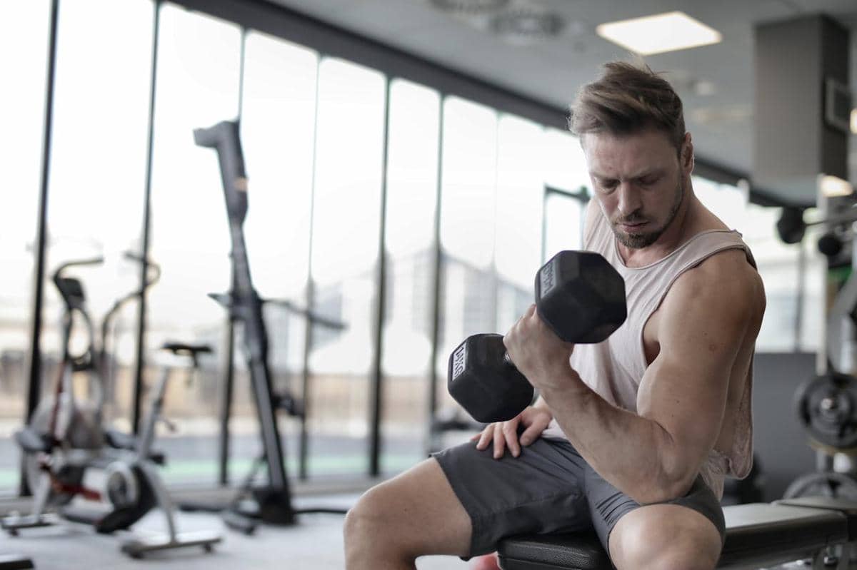 Seorang pria berolahraga di gym sambil mengangkat dumbel dengan satu tangan, mengenakan kaus tanpa lengan dan celana pendek.
