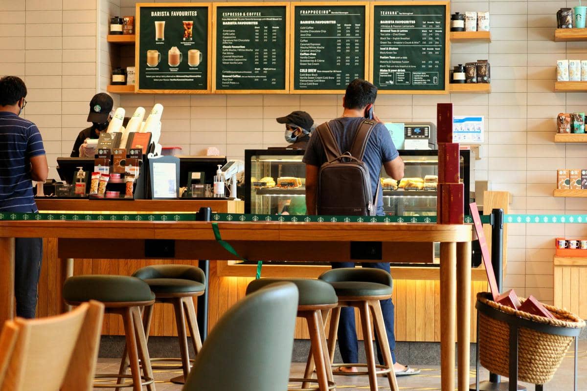 Suasana di dalam coffee shop modern dengan pelanggan sedang memesan minuman di kasir dan barista menyiapkan pesanan di balik meja.