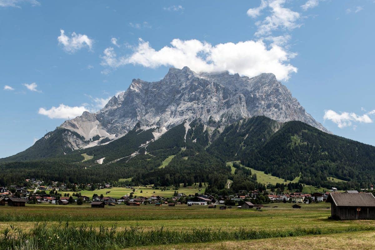 Pemandangan gunung Zugspitze dengan puncak berbatu menjulang di bawah langit biru, dikelilingi hutan hijau dan desa di lembah.