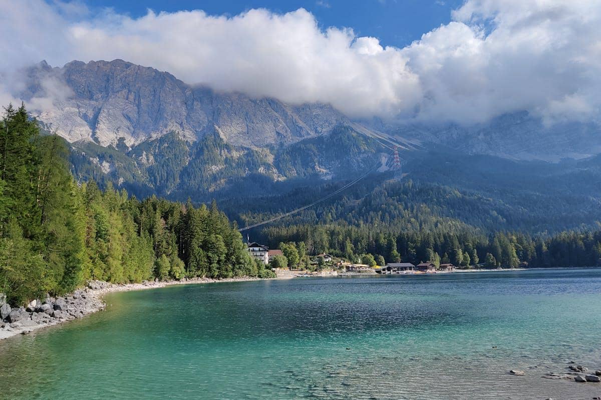 Pemandangan gunung Zugspitze dengan hutan hijau di tepi danau berair jernih berwarna biru kehijauan di bawah langit berawan.