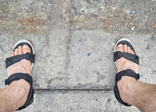 Kaki seseorang mengenakan sandal hitam dengan tali di atas permukaan jalan beton berwarna abu-abu.