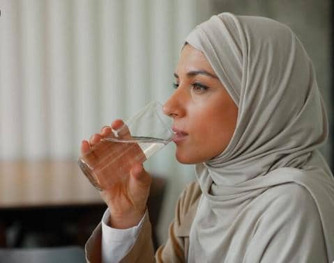 Seorang wanita berhijab duduk sambil meminum segelas air putih di dalam ruangan dengan latar belakang lembut.