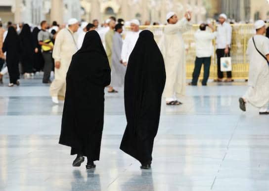 Dua perempuan mengenakan pakaian hitam berjalan kaki di area terbuka dengan banyak orang berpakaian putih di sekitarnya.