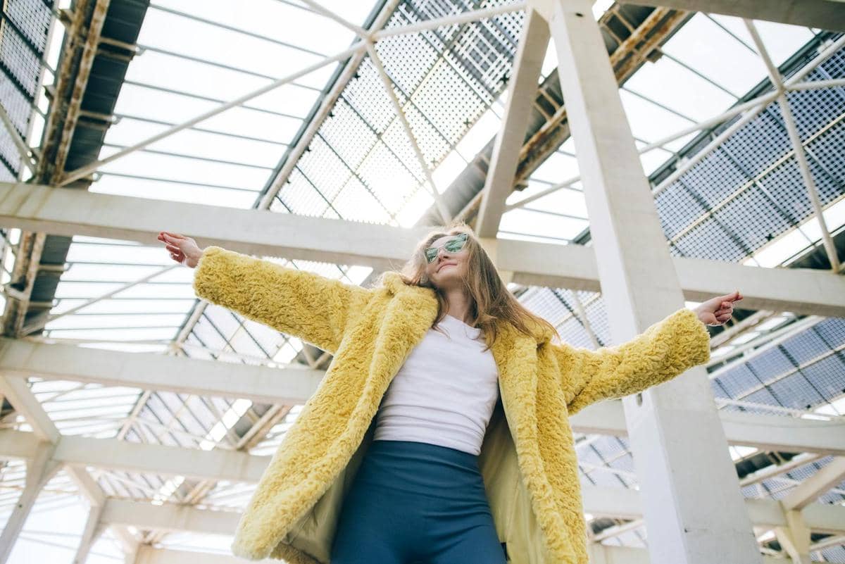 Seorang perempuan mengenakan mantel bulu kuning dan kacamata hitam berdiri dengan tangan terbuka di bawah struktur baja beratap kaca.