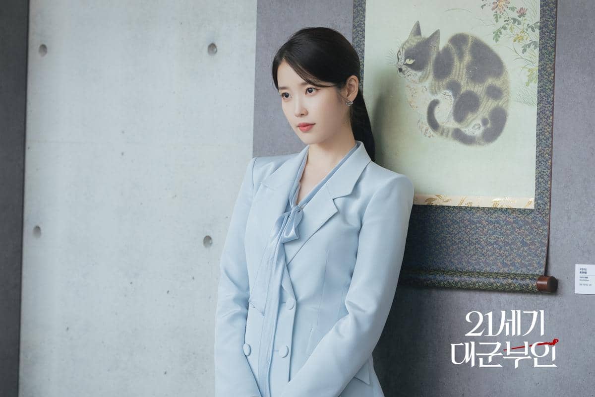 Seorang wanita mengenakan setelan jas biru muda berdiri di depan dinding abu-abu dengan lukisan kucing di belakangnya.